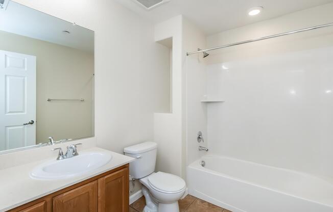A clean, modern bathroom featuring a white bathtub with a shower, a toilet, and a vanity with a sink and mirror. The walls are painted light colors, and there are tiled floors. The overall design is simple and spacious, with ample natural light.
