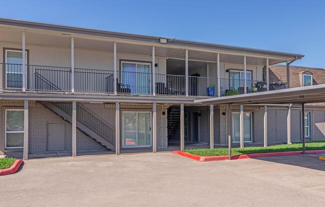 Two-story apartment building with a covered parking area in front. The building features multiple balconies, a staircase on the left side, and large windows. The surrounding area has green grass and a clear blue sky, indicating a bright and sunny day.