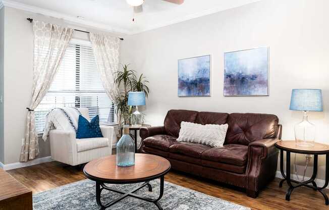 A living room with a brown leather couch, a white chair, a wooden coffee table, and two paintings on the wall.