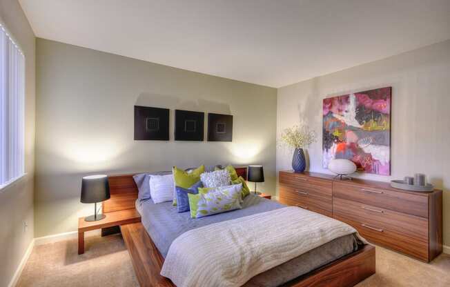 Large Bedroom with Full Sized Bed on Elevated Wood Frame, Carpet, Wood Dressers, Small Black Lamps on each side of Bed, Abstract Painting on Wall