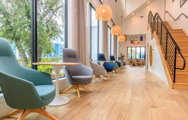 A bright and modern interior featuring a hallway with large windows, wooden flooring, and stylish hanging light fixtures. Comfortable chairs in varying shades of blue and grey are paired with small tables, while a staircase with a black railing leads to an upper level, creating a welcoming atmosphere.