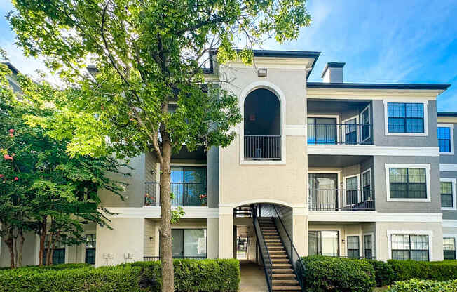 A three-story apartment building at Saxony at Chase Oaks Apartments in Dallas, TX, surrounded by a green lawn, clear blue skies, and neatly trimmed shrubs.