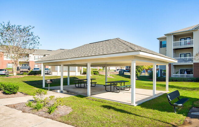 A covered picnic area with benches and a table is surrounded by a grassy area.