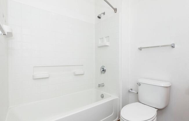 A clean, modern bathroom featuring a white bathtub with a showerhead, a toilet, and white tiled walls. The space is well-lit, giving it a fresh and bright appearance, with minimal decor and a simple design.