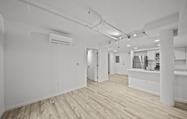 Modern empty room with light wood flooring, white walls, and a column. A doorway leads to a kitchen equipped with stainless steel appliances, visible in the background. Soft lighting fixtures are installed on the ceiling, and an air conditioning unit is mounted on the wall.