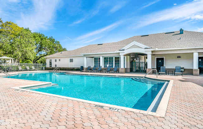 Swimming pool with large tanning deck, table with umbrella, lounge chairs, clubhouse in background surrounded by native landscaping