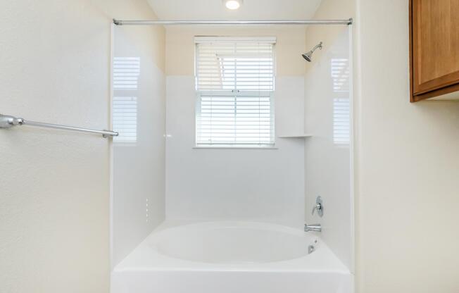 A clean and modern bathroom featuring a bathtub with a shower, surrounded by white walls. Natural light enters through a window with blinds. There is a wooden cabinet on the right side of the image. The overall aesthetic is bright and minimalistic.