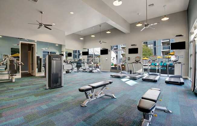 A spacious fitness center here at Rio Paradiso featuring a wide open layout with treadmills, stationary bikes, weight machines, and adjustable benches arranged on patterned flooring, complemented by high ceilings with ceiling fans, wall-mounted televisions, and large windows that bring in natural light and views of the outdoor pool area.