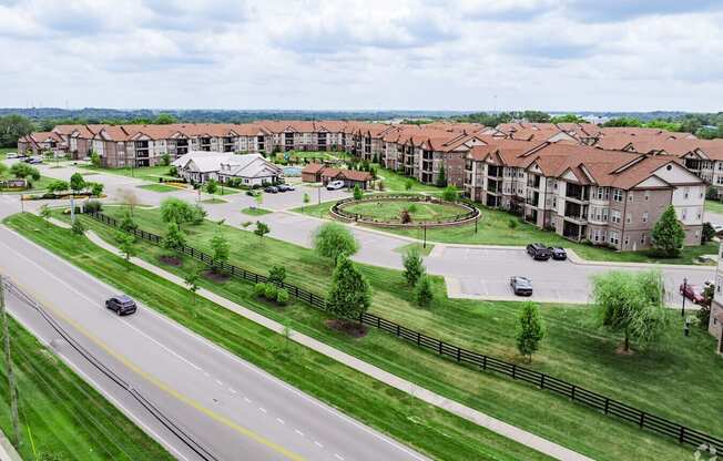 A road with cars and a grassy area with trees and apartment buildings in the background.