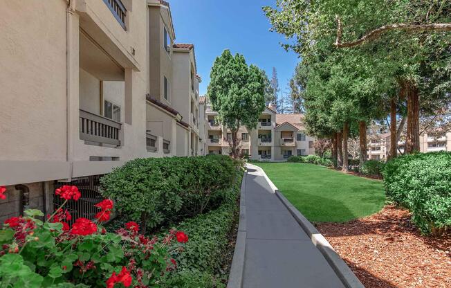 A landscaped path lined with vibrant red flowers leads through a residential area. The path is flanked by greenery and trees, with beige apartment buildings visible in the background under a clear blue sky. The scene conveys a peaceful, inviting atmosphere.