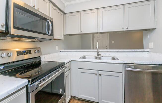 Modern kitchen featuring stainless steel appliances, including a microwave, stove, and dishwasher. The cabinetry is a light gray color, complemented by a white countertop with a marbled design. A double sink is positioned beneath a mirror, enhancing the space's openness. Wood-like flooring completes the contemporary look.