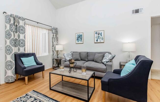 A modern living room featuring a gray sofa with decorative pillows, two blue accent chairs, and a coffee table with decorative items. Light-colored curtains frame a window, and wall art adds a touch of style. Warm wooden flooring complements the cozy atmosphere.