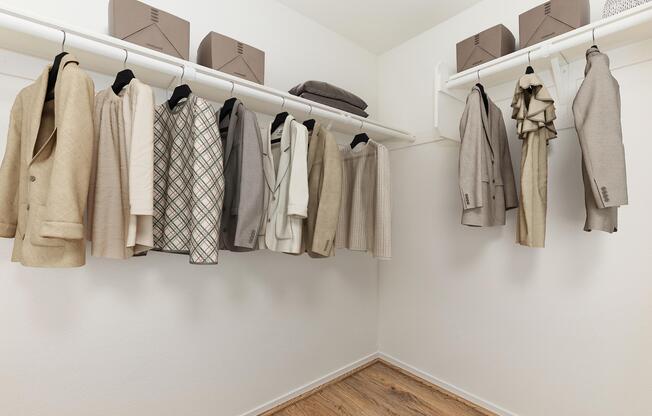 A neatly organized closet featuring various neutral-toned jackets and blazers hanging on a white rack. Above the clothing, there are storage boxes, and the floor is made of light wooden planks, creating a clean and modern aesthetic.