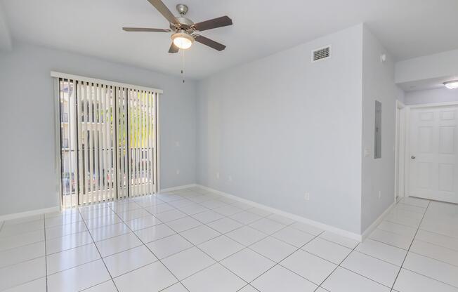 A spacious living area with light gray walls and large white tiles on the floor. A ceiling fan is visible, and there are sliding glass doors with vertical blinds that open to an outdoor space. The room is bright and empty, with a door leading to another area in the background.