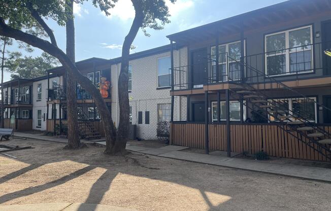 Row of modern two-story apartment buildings with balconies, surrounded by trees. A sandy area in the foreground, and blue sky with scattered clouds above. The setting appears inviting with outdoor seating and greenery.