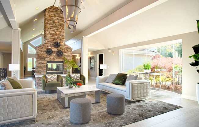 A spacious resident lounge here at Andorra featuring a dramatic stone fireplace with a vaulted ceiling, modern pendant lighting, plush sofas and accent chairs arranged around a sleek coffee table, soft patterned carpeting, and large windows that fill the space with natural light and offer views of landscaped greenery.