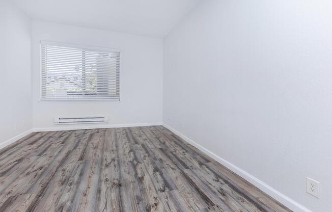 A bright, vacant room with light gray walls, a window featuring white blinds, and modern wood-like flooring. The room has minimal furniture, creating an open and spacious feel, with natural light coming in from the window.
