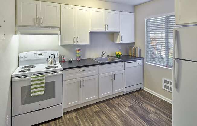 Kitchen with white cabinets and grey countertops. Image contains a white oven, sink and white dishwasher. One window and wood style flooring.at Woodhaven, Everett, Washington