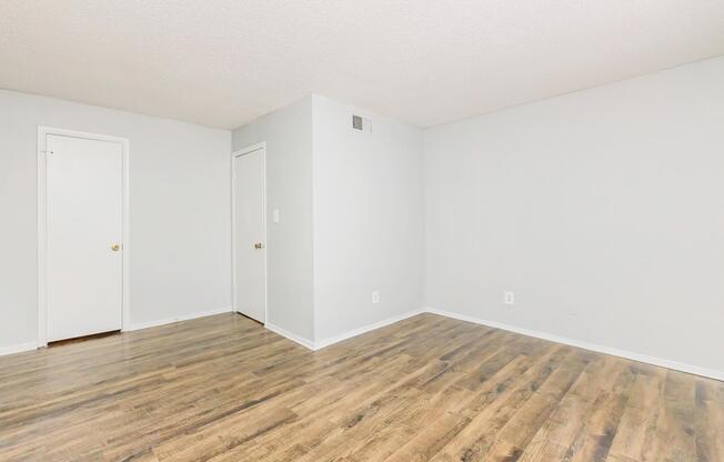 Empty room with light gray walls and two doors (one closed) on the left side. The floor is wooden with a warm tone. There are no furnishings or decorations, creating a spacious and minimalist appearance. The lighting is bright, contributing to an open and airy feel.
