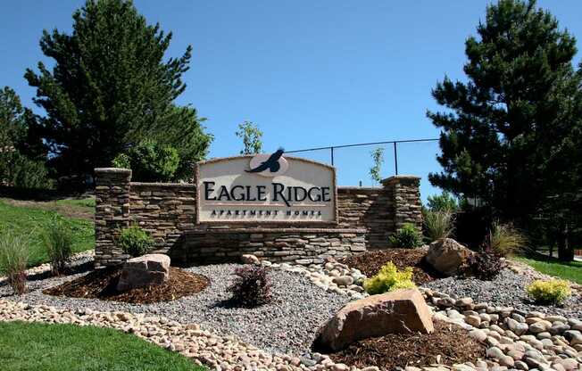 A prominent stone monument sign here at Eagle Ridge set within manicured landscaping, featuring layered stonework, decorative rock beds, low shrubs, and large accent boulders, framed by tall evergreen trees and a gently sloped grassy hillside beneath a clear blue sky.