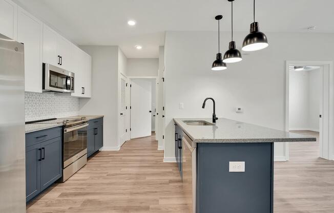 Modern kitchen with stainless steel appliances, including a microwave and oven, grey countertops, and blue cabinets. Features pendant lighting above a large island, light-colored walls, and wood flooring. The room connects to other spaces with white doors and a clean, open layout.