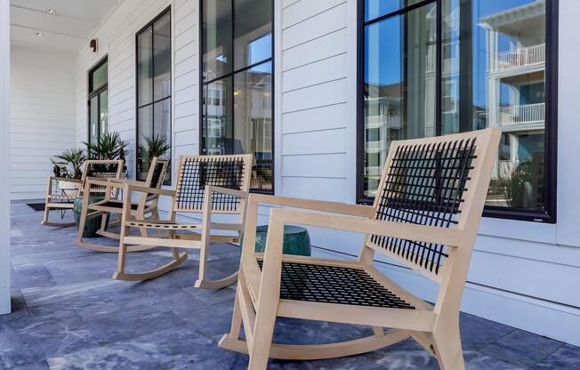A row of modern wooden rocking chairs with black woven seats, placed on a stone-tiled porch. Large windows with reflections of nearby buildings and plants enhance the serene outdoor setting.