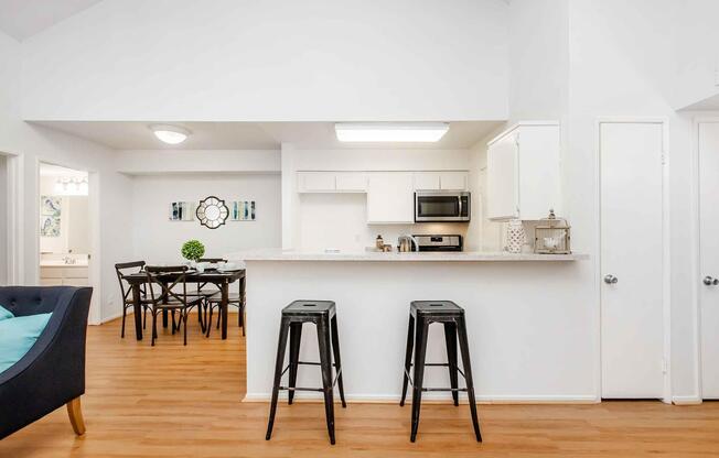 A bright, modern kitchen with white cabinets, granite countertops, and stainless steel appliances. In the background, there is a dining area with a black table and chairs. Two black bar stools are placed in front of the kitchen counter. The space features wooden flooring and a simple, clean aesthetic.