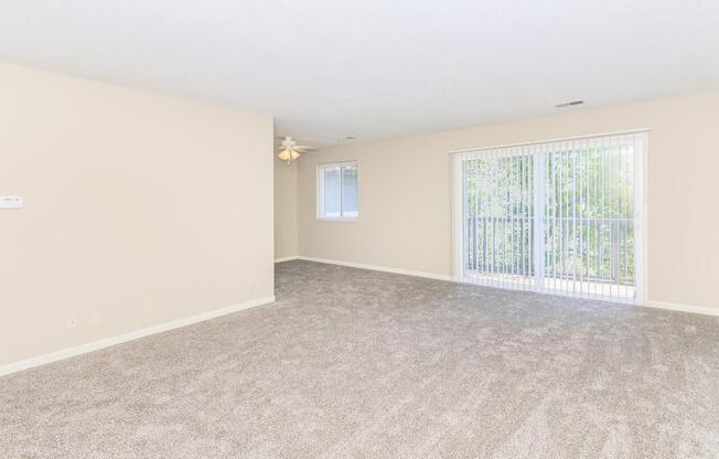 Spacious empty living room featuring light beige walls, a ceiling fan, and soft carpet. Large sliding glass door leads to a balcony with greenery visible outside. Natural light fills the room, creating a bright and airy atmosphere.