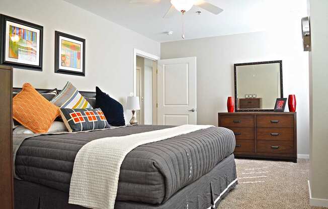 bedroom with light carpeting, bed with gray duvet, brown dresser with mirror at Residences at Forest Park, Saint Louis, MO, 63108