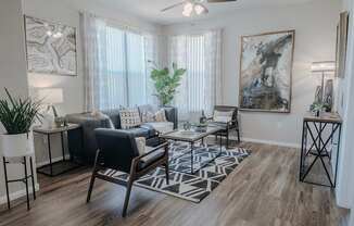 A living room with a black and white rug, a black leather couch, a wooden chair, a white side table, a potted plant, and a painting on the wall.