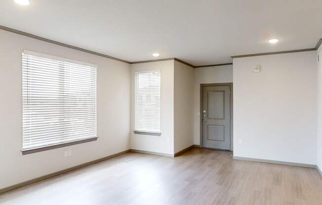 A modern, empty living room here at 49 West Apartments with light wood flooring, white walls, and two large windows with blinds. A closed gray door is on the right. Bright and minimalist.