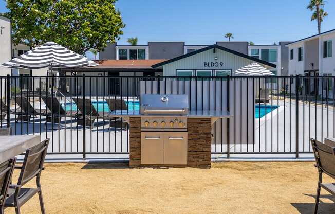 A BBQ grill is in the middle of a sandy area with a pool and umbrellas in the background.