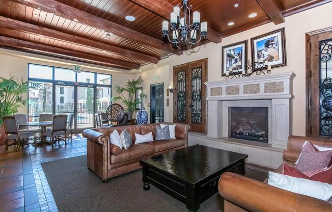 A cozy lounge area featuring a leather sofa and armchair, a wooden coffee table, a decorative fireplace, and potted plants. The room has wooden beams on the ceiling, framed artwork on the walls, and large windows that provide natural light, creating an inviting atmosphere.