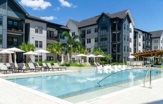 A swimming pool surrounded by lounge chairs and umbrellas in front of a row of apartment buildings.