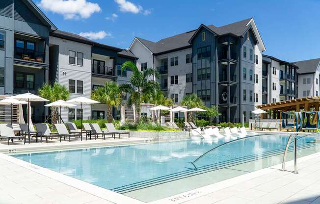 A swimming pool surrounded by lounge chairs and umbrellas in front of a row of apartment buildings.
