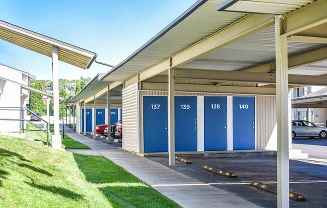 Covered parking and storage units with blue doors next to sidewalk lined with green grass in Pocatello, ID