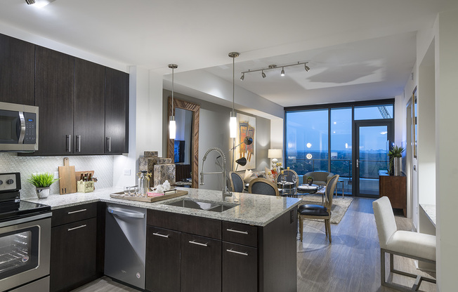 Open-concept kitchen, dining, and living space with ash-gray wood-style floors, dark modern cabinets, stainless steel appliances, granite countertops, and a spacious living area with a full-width floor-to-ceiling window.