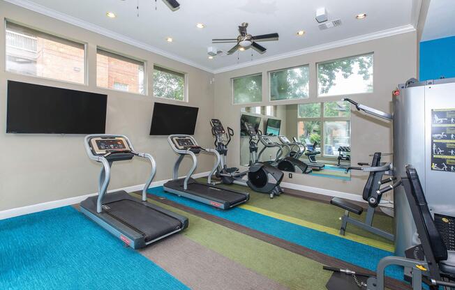A modern fitness room featuring two treadmills, a stationary bike, and various exercise equipment. The space is well-lit with natural light from large windows and has a colorful, patterned carpet. Two flat-screen TVs are mounted on the wall for entertainment during workouts.
