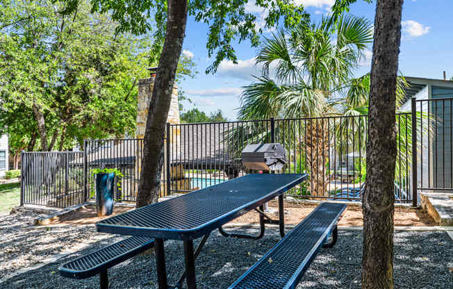 two picnic tables and a grill in a fenced in area with trees