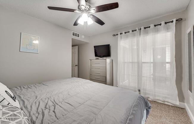 A cozy bedroom featuring a light gray wall, a queen-sized bed with a gray comforter, and a ceiling fan. There's a tall dresser against one wall, a flat-screen TV mounted on another, and large windows with sheer curtains allowing natural light to fill the room. A framed artwork is visible on the wall.
