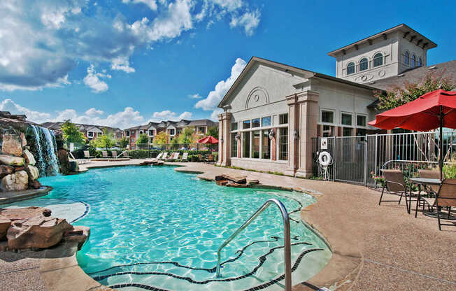 A resort-style pool here at Landing at Mansfield with curved entry steps, rock waterfall feature, turquoise water, tan pool deck, red patio umbrellas, lounge chairs, and a bright clubhouse building under a blue sky.