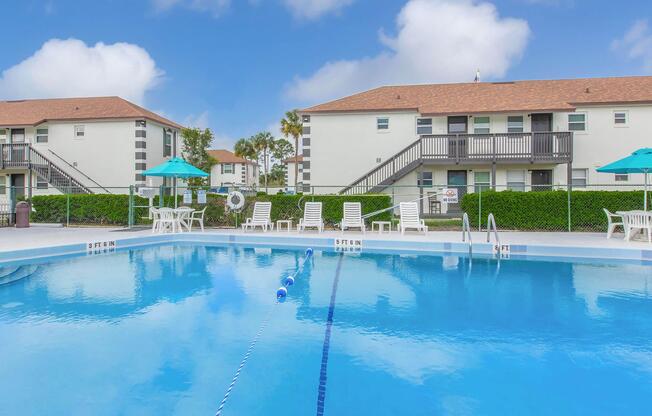 A clear blue swimming pool surrounded by lounge chairs and umbrellas, with two apartment buildings in the background. The sky is bright with fluffy white clouds, and the pool area is well-maintained, featuring diving lanes and a landscaped border.