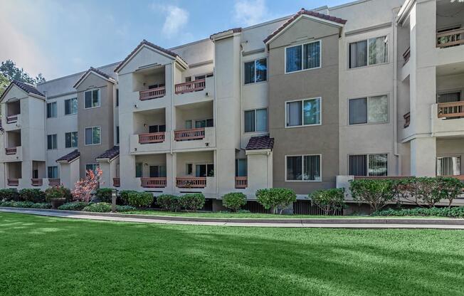 A modern apartment building with multiple floors, featuring balconies and large windows. The exterior is painted in light beige with dark accents. In front, there is a well-maintained green lawn and landscaped shrubs, creating a pleasant outdoor space. The scene is bright and inviting.