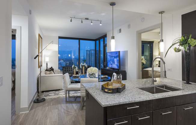 Open-concept kitchen, dining, and living space with 10-ft floor-to-ceiling windows on two walls, ash-gray wood-style floors, dark modern cabinets, stainless steel appliances, granite countertops, and a spacious living area.