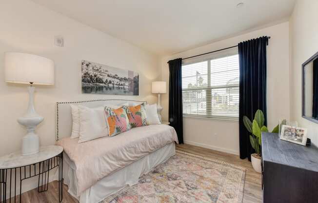 Bedroom with hardwood inspired flooring, a day bed against the wall, table with lamp, dresser and large window with darker curtains at Morgan Ranch Apartments, Morgan Hill, CA