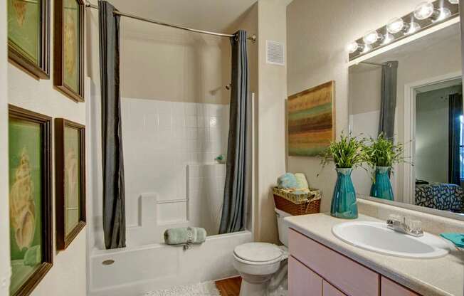 A bright and well-appointed bathroom here at Pala Mesa featuring a full tub and shower combination with tile surround and a curved curtain rod. A spacious vanity with ample counter space and a large mirror is complemented by modern lighting, while decorative accents and soft towels create a clean and inviting atmosphere.