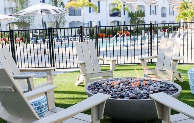 a fire pit with chairs and umbrellas on a lawn with a pool in the background