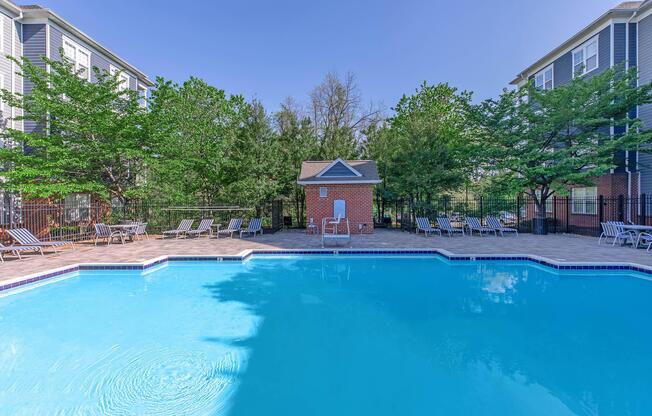A clear blue swimming pool surrounded by lounge chairs and green trees. In the background, there are two multi-story buildings. A small brick building is visible near the pool, likely serving as a pool house or storage area. The sky is bright and clear, indicating a sunny day.