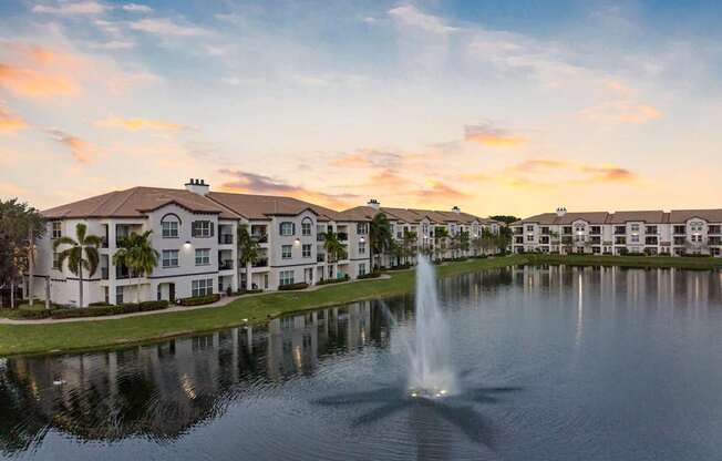 A fountain in the foreground of a serene lake in front of apartment buildings.