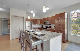 A spacious kitchen here at Vue at Pinnacle Park with warm wood cabinets, granite countertops, stainless steel appliances, and a large island with bar seating, pendant lighting, and a bright window for natural light.
